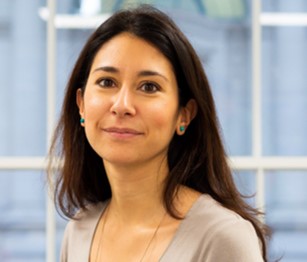 A person with long, dark hair wearing a light-colored top, standing indoors in front of large windows. The face is blurred for privacy.