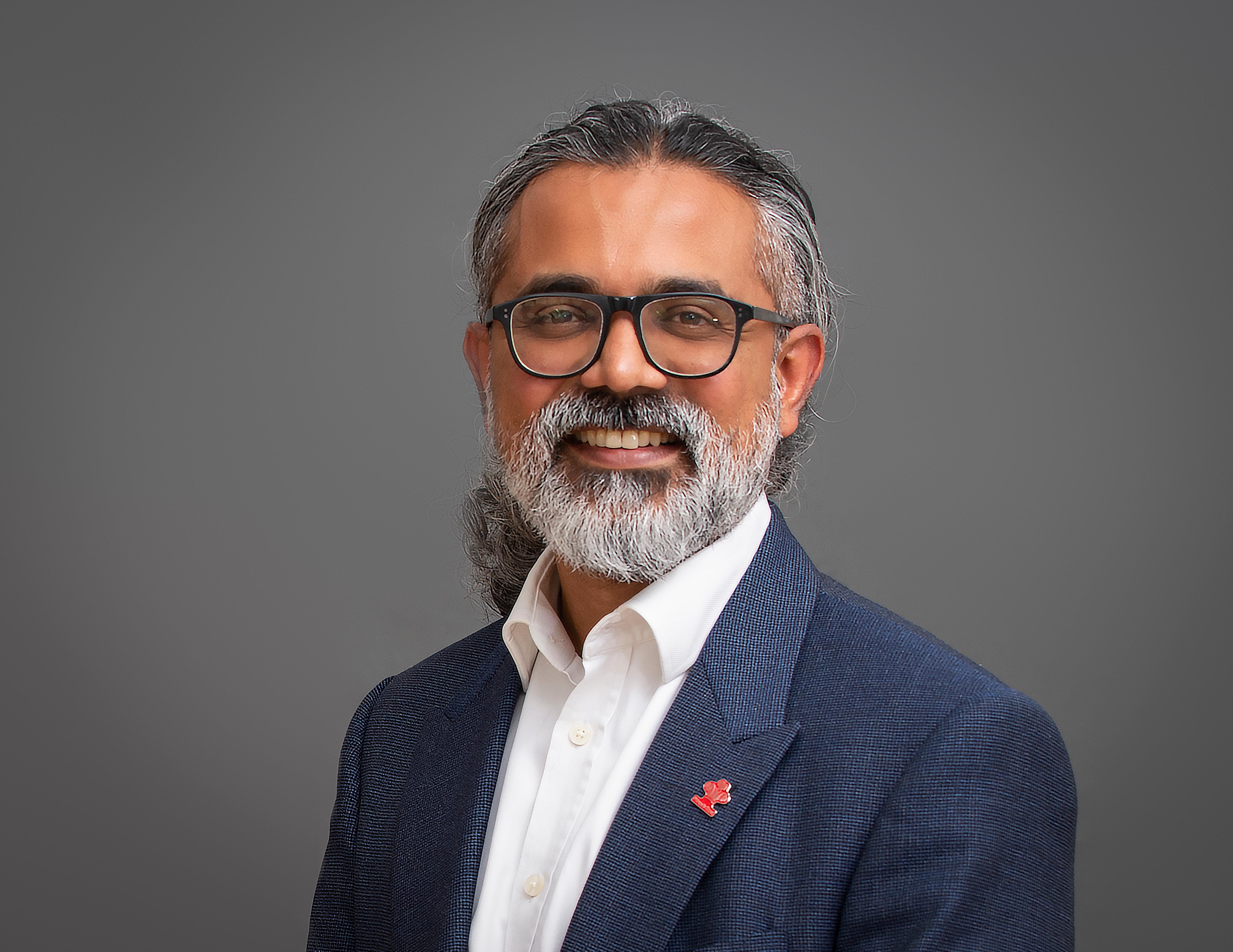 Person with blurred face in white shirt and blue blazer with a red pin, standing against a gray background.