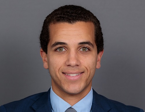 Person in a blue suit and tie with a blurred face, standing against a gray background.
