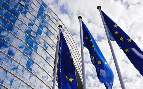 Three EU flags waving in front of a modern glass building with a partly cloudy sky reflected in the windows.