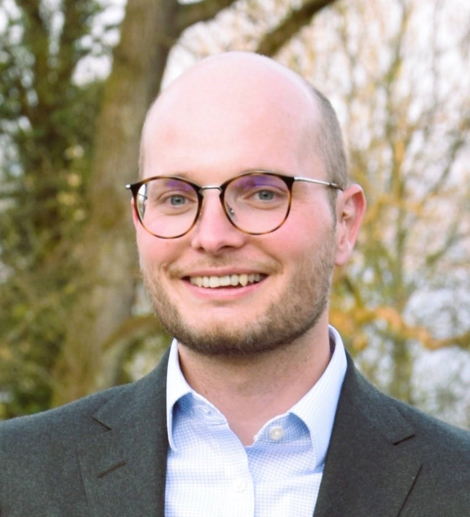 Person in a suit and light blue shirt with a blurred face, standing in front of trees.