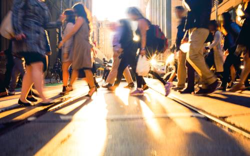 Street-level view of a busy urban crosswalk with people walking, sunlight casting long shadows and a warm glow on the pavement.