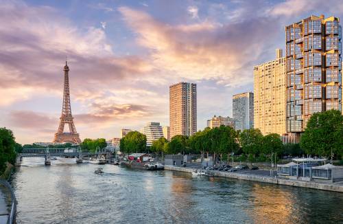 View of the Seine River in Paris with the Eiffel Tower, modern buildings, and boats under a glowing sky.