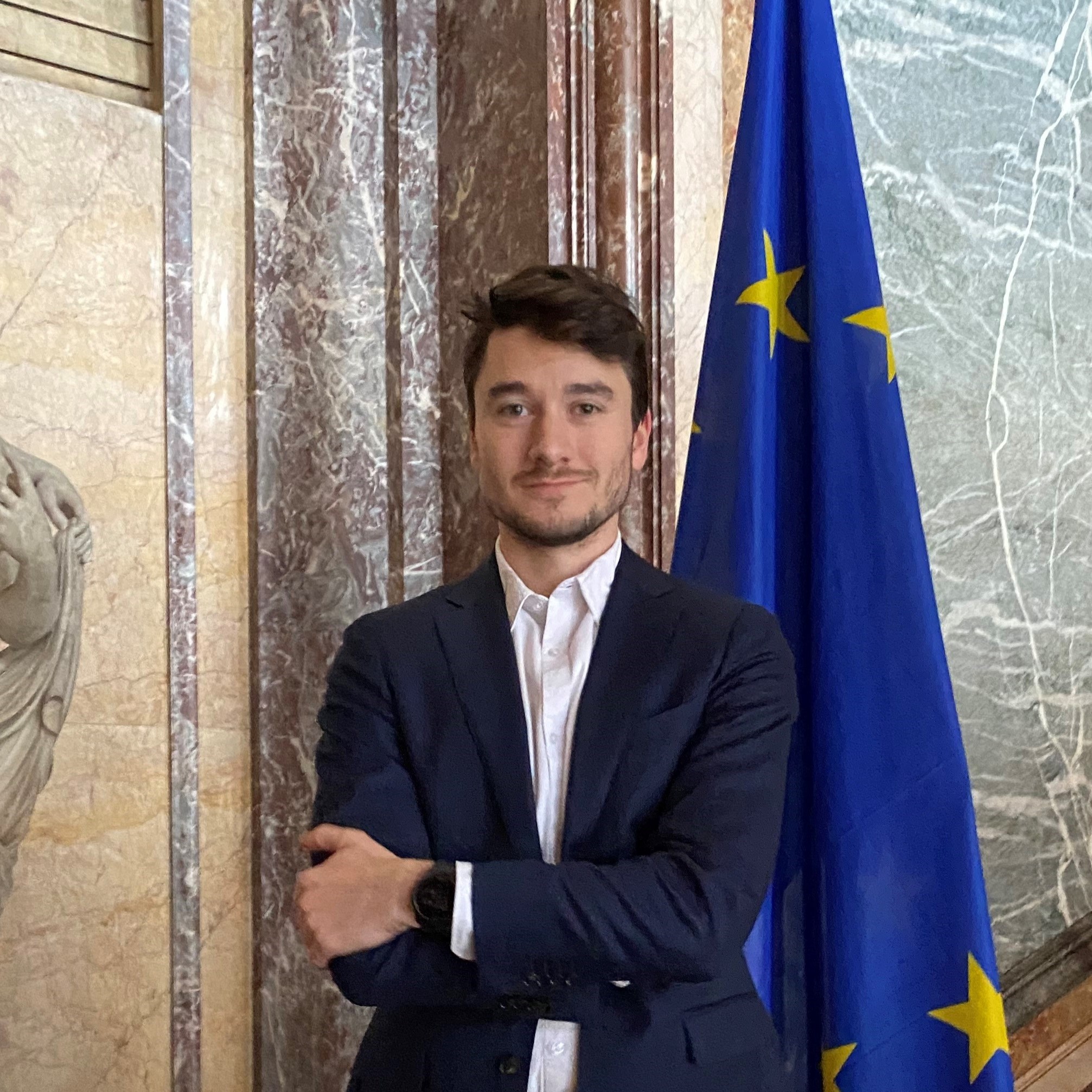 A well-dressed man in a suit and tie poses in front of the European flag, representing international relations.