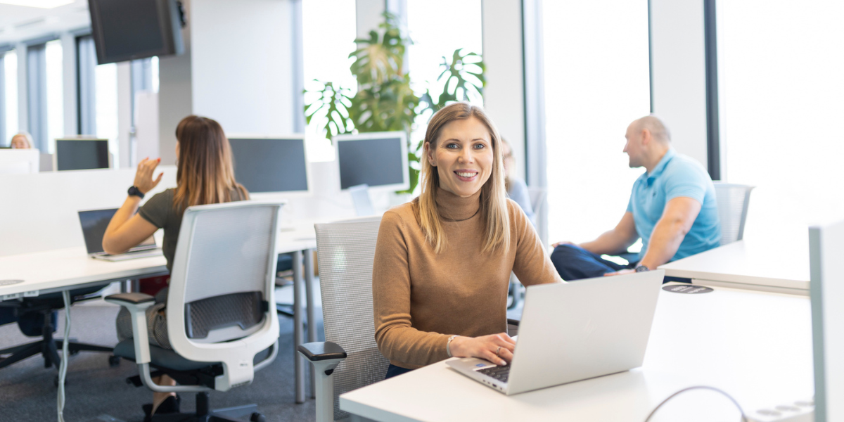 młoda kobieta uśmiechająca się na wprost kamery, siedzii na open space przy laptopie