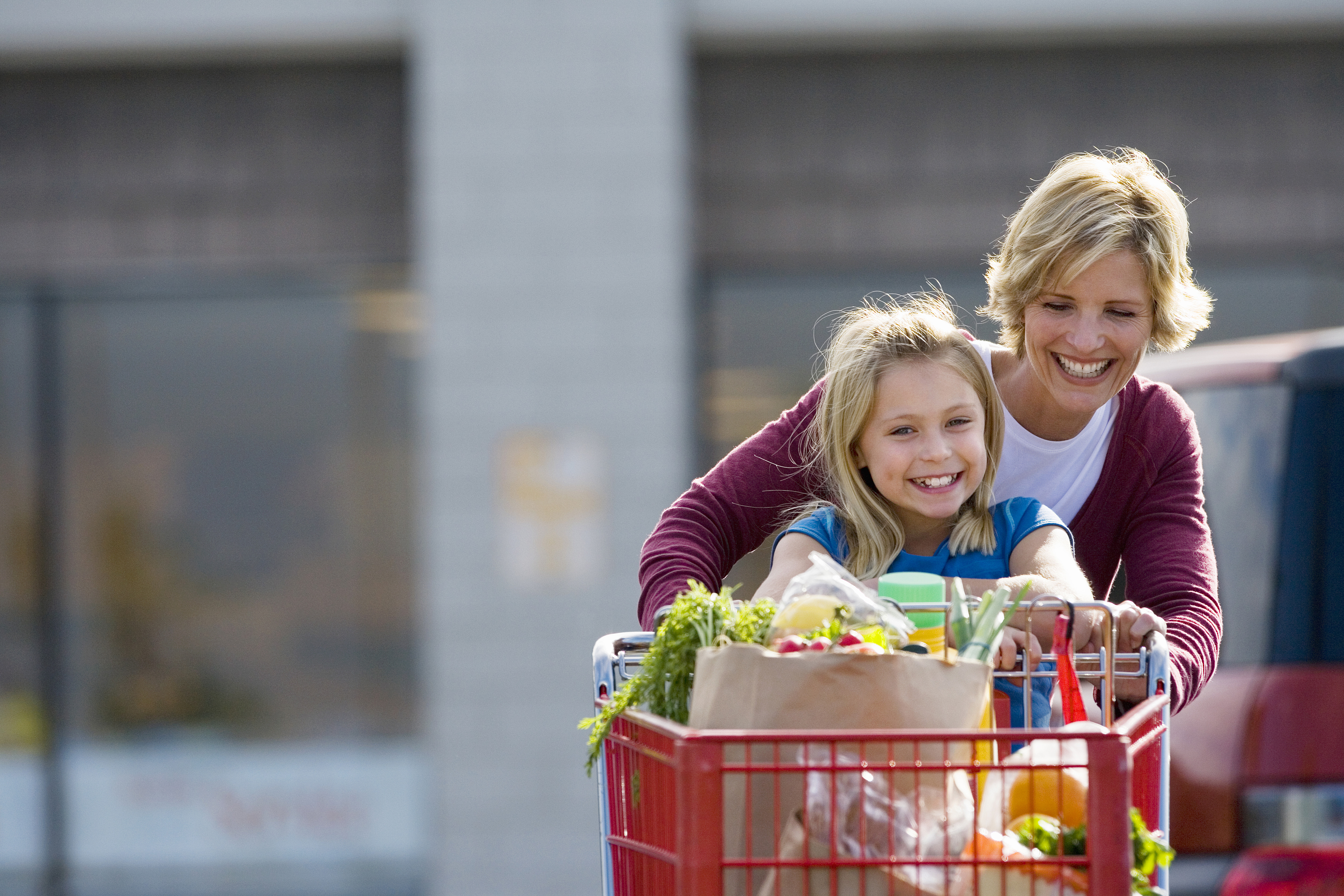 Mother and daughter pushing shopping trolley in supermarket car park, smiling, front view, portrait