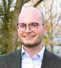 Person in a suit and light blue shirt with a blurred face, standing in front of trees.