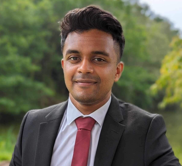 A man in a suit and tie stands confidently by a river, with water flowing gently in the background.